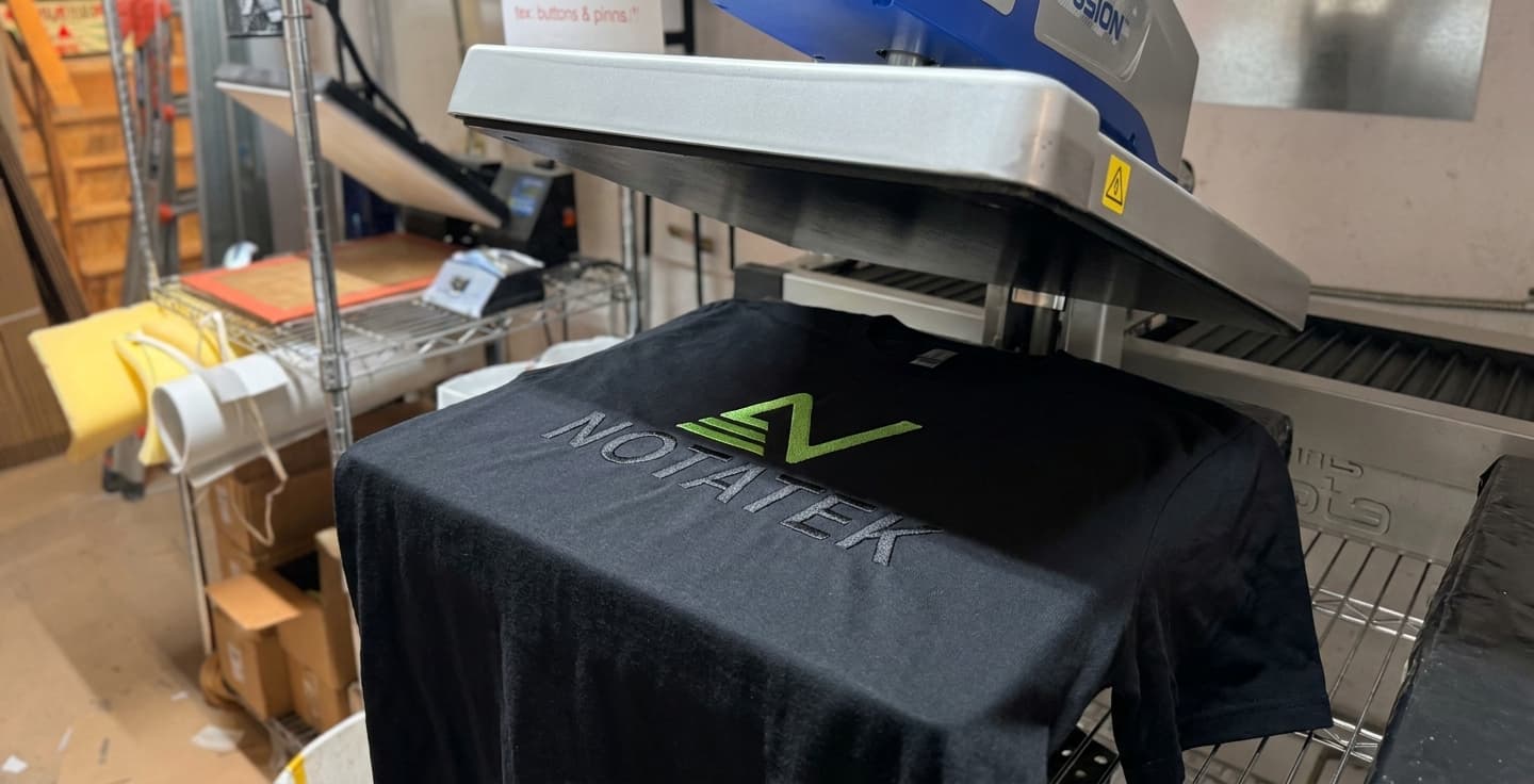 Black t-shirt with a green and grey NOTATEK logo on a heat press machine during production.