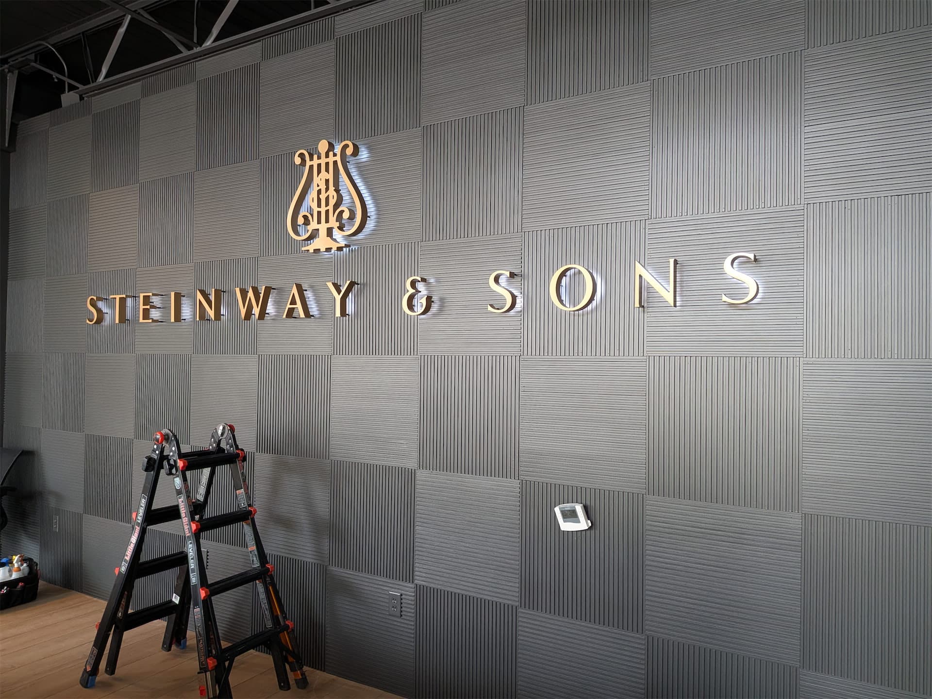 Gold illuminated Steinway & Sons logo and lettering mounted on a grey textured wall.