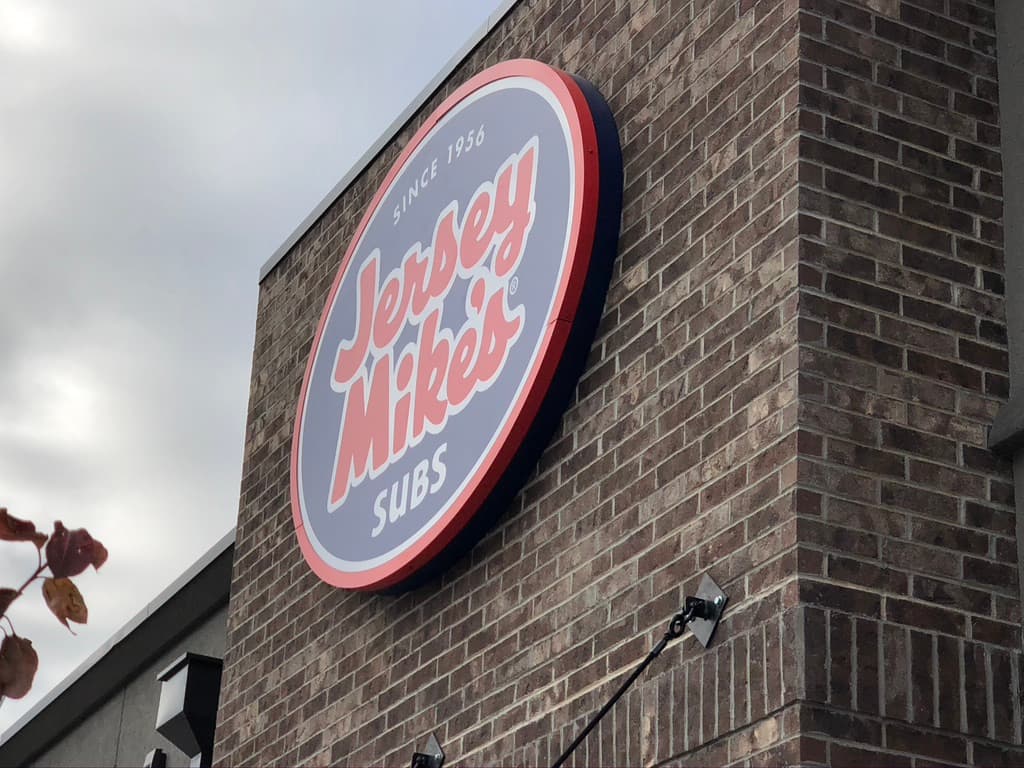 A round, illuminated Jersey Mike's Subs sign mounted on a brown brick exterior wall.