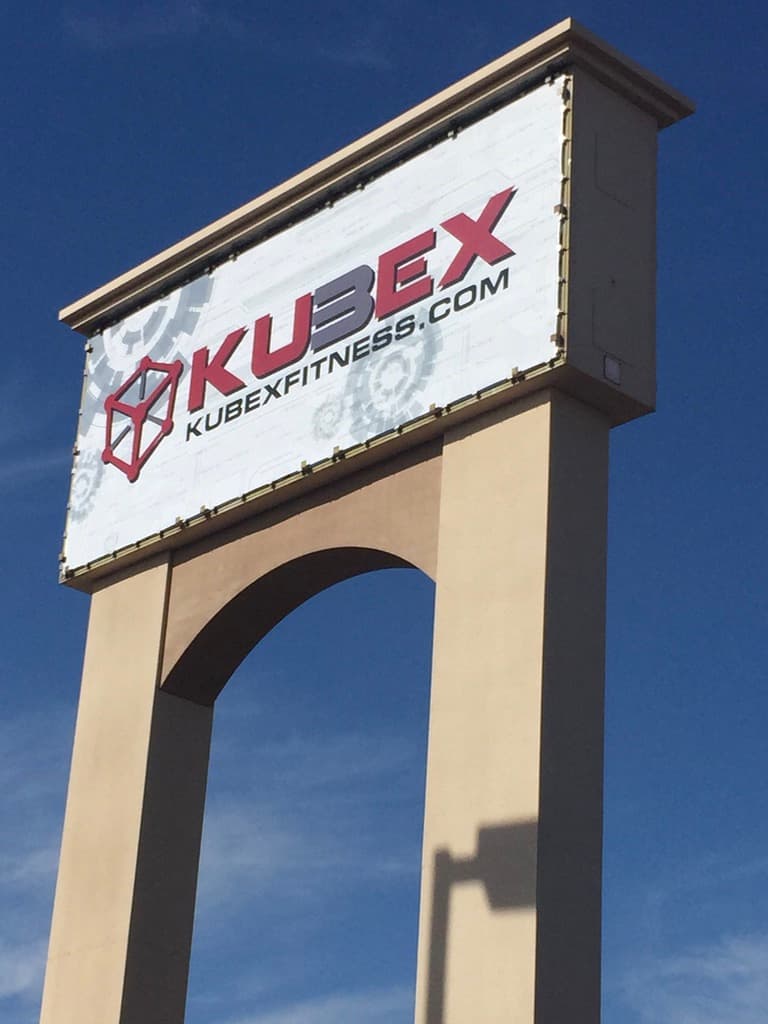 Outdoor pylon sign for Kubex Fitness featuring their red and black logo against a blue sky.