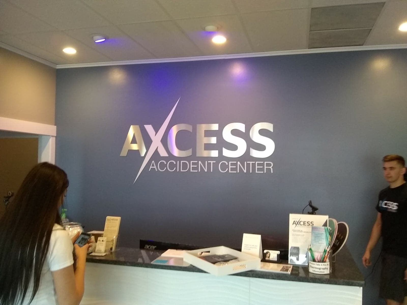 Brushed metal dimensional lobby sign for Axcess Accident Center mounted on a blue reception wall.