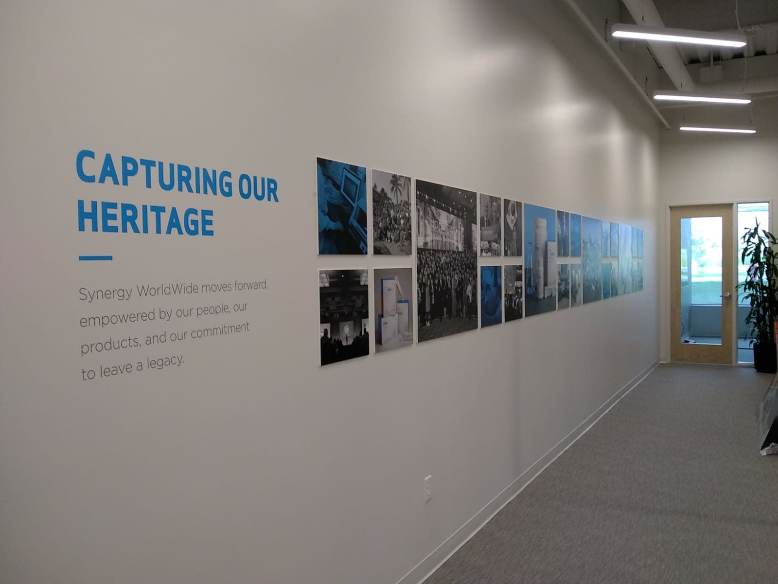 Office hallway featuring blue wall graphics reading 'Capturing Our Heritage' alongside a photo collage wall display.