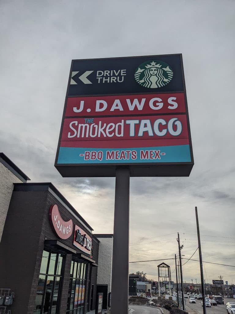 A tall outdoor pylon sign displaying logos for Starbucks Drive Thru, J. Dawgs, and The Smoked Taco.
