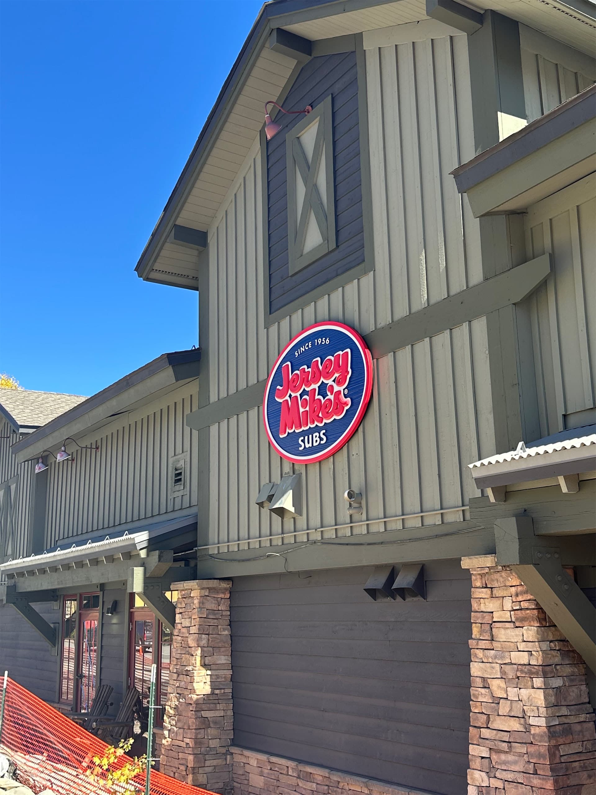 A circular Jersey Mike's Subs illuminated sign mounted on a grey wooden exterior wall.