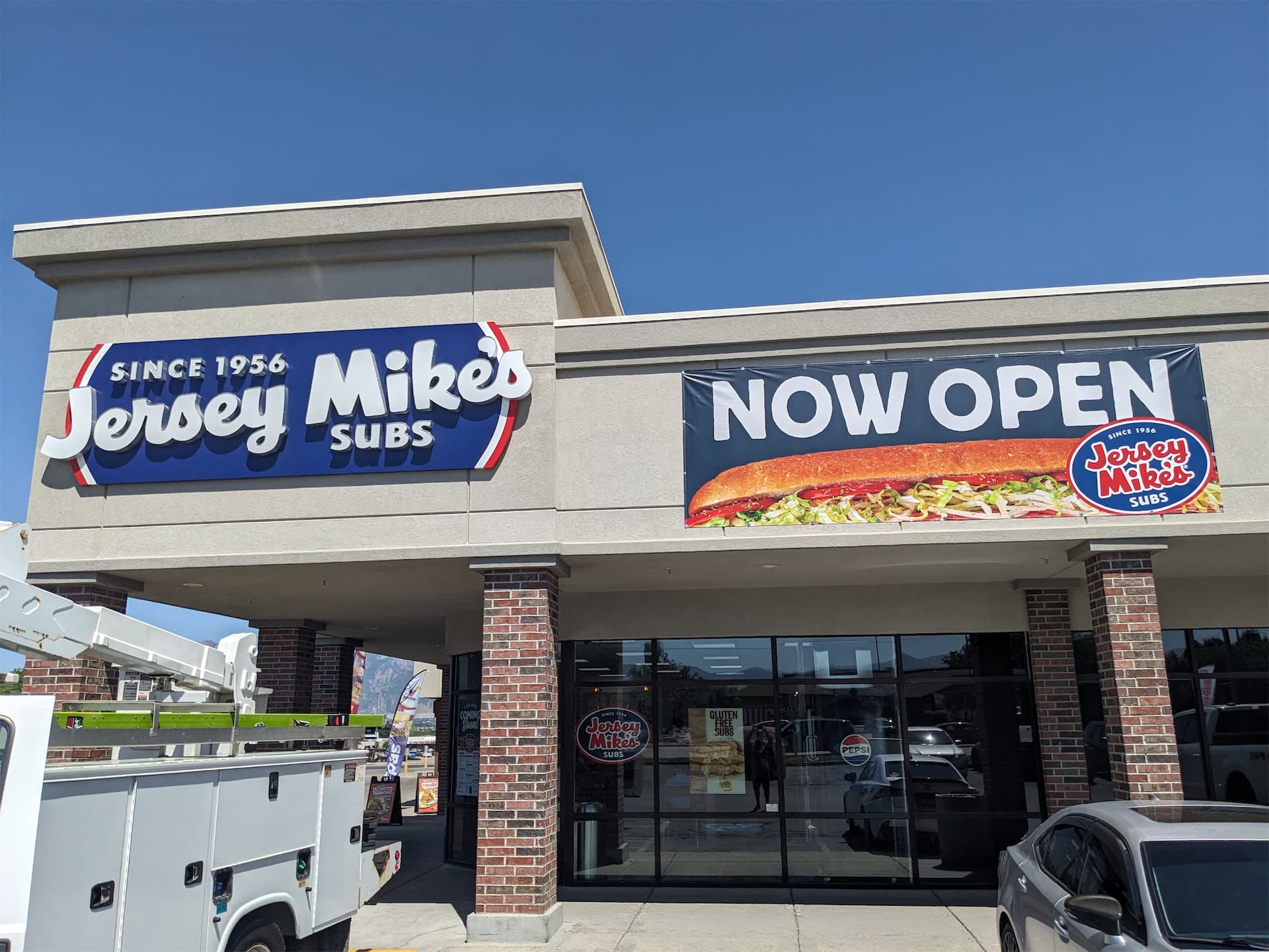 Exterior storefront of Jersey Mike's Subs featuring illuminated building signage and a Now Open banner.