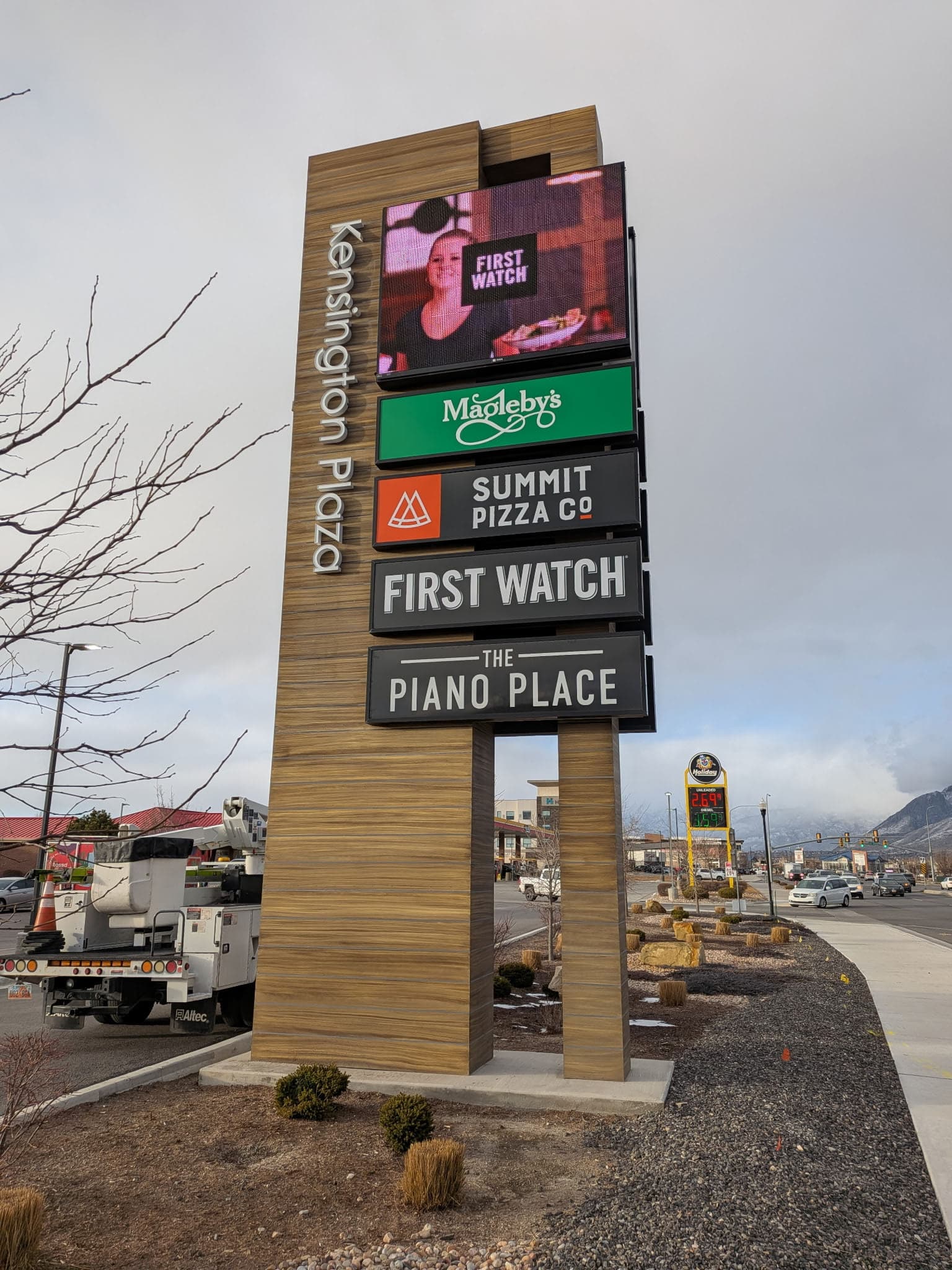 Tall wood-textured pylon sign for Kensington Plaza showcasing multiple tenant logos including Summit Pizza.