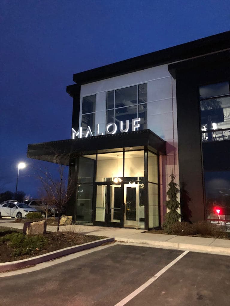 Illuminated white MALOUF sign mounted above a modern office glass entrance at dusk.