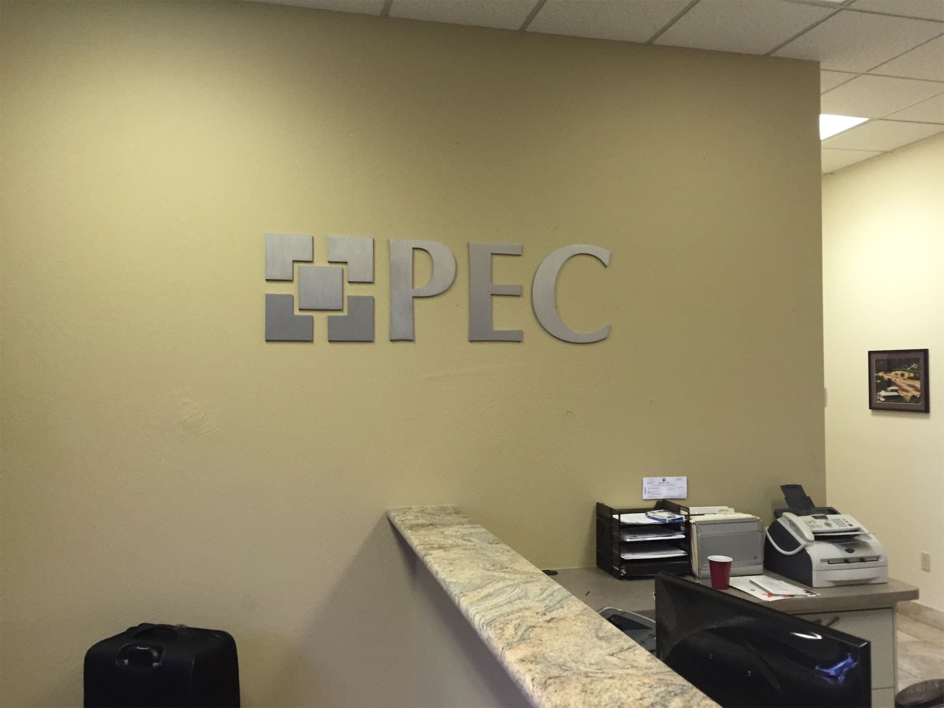 Brushed metal dimensional PEC logo sign mounted on an office wall behind a reception desk.