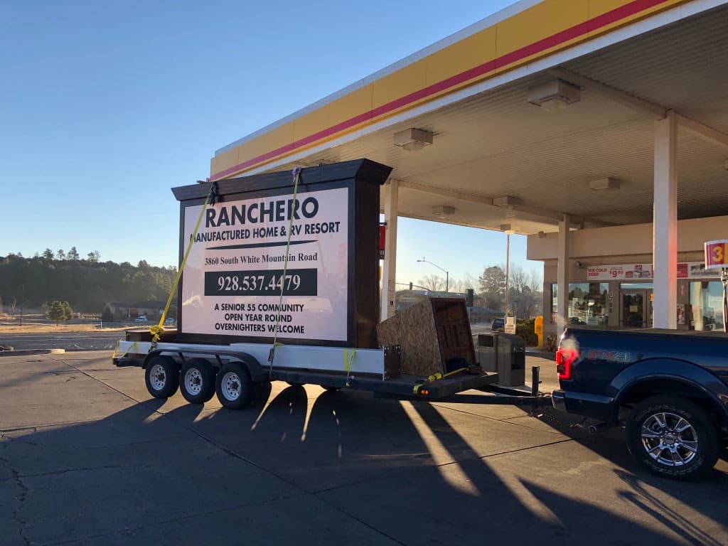 A large Ranchero Manufactured Home & RV Resort sign secured on a flatbed trailer towed by a pickup truck.