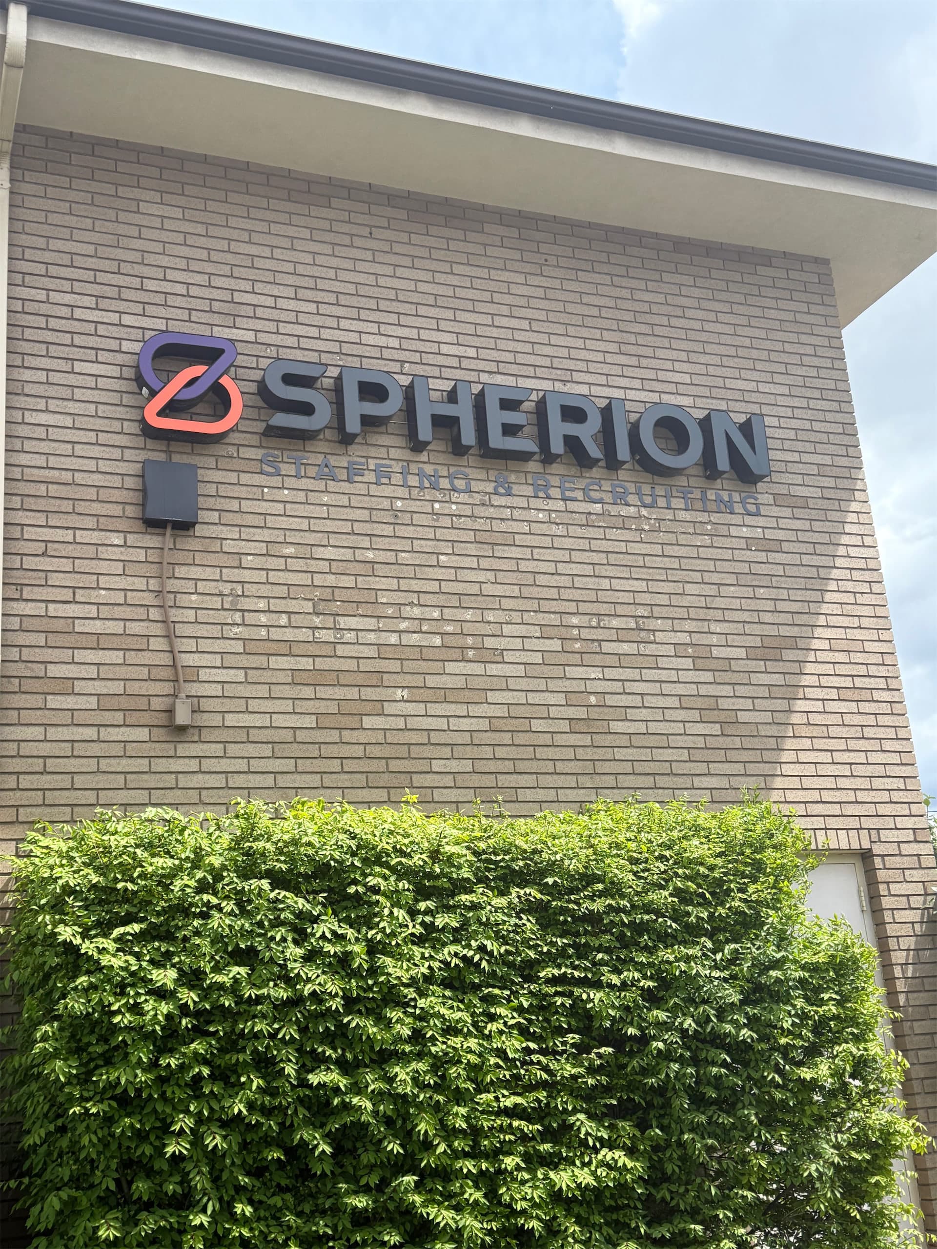 Spherion Staffing & Recruiting dimensional letter sign mounted on a brick building exterior behind green foliage.