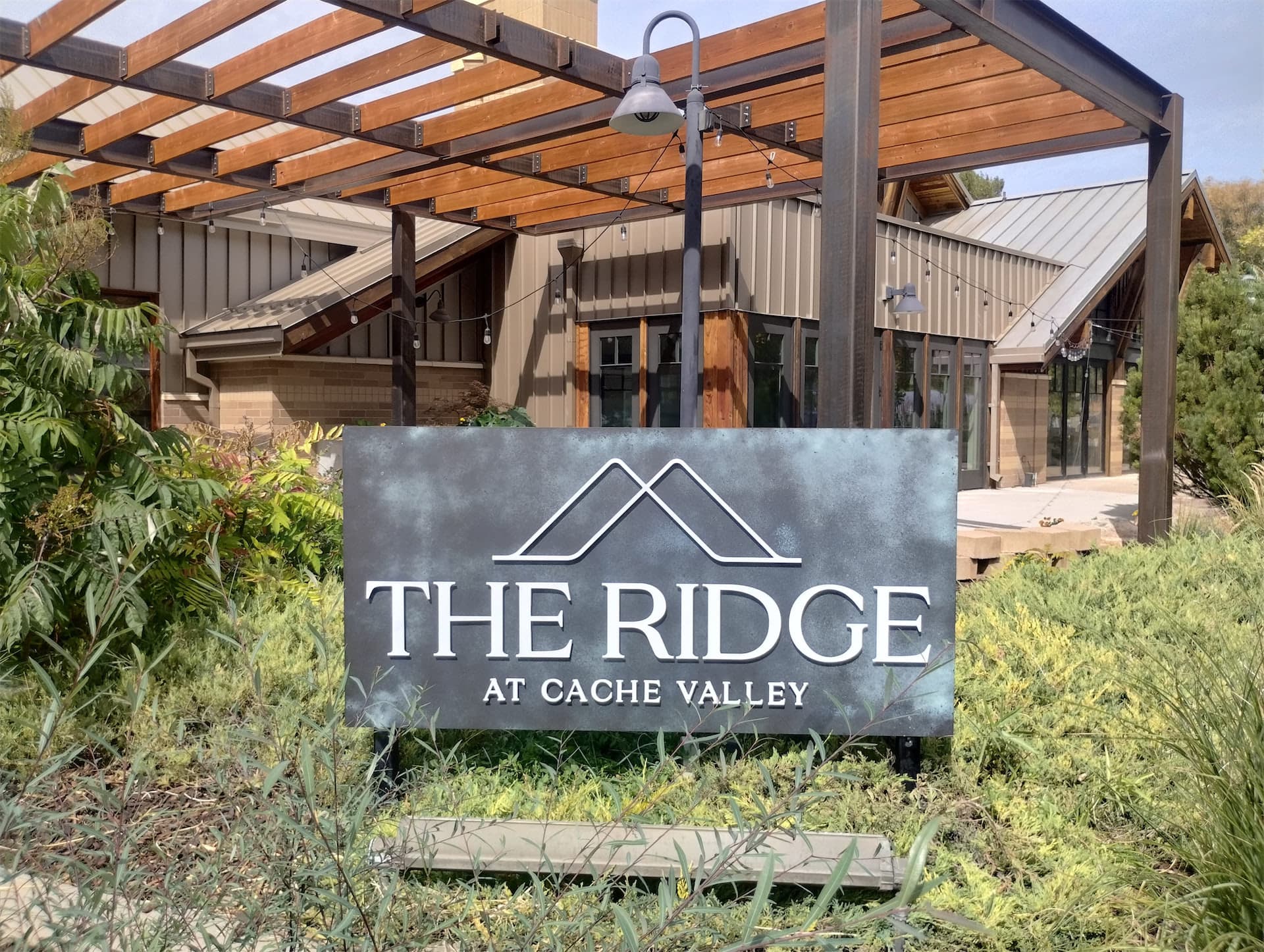 Exterior sign for The Ridge at Cache Valley with a metal finish and white logo in front of a building.