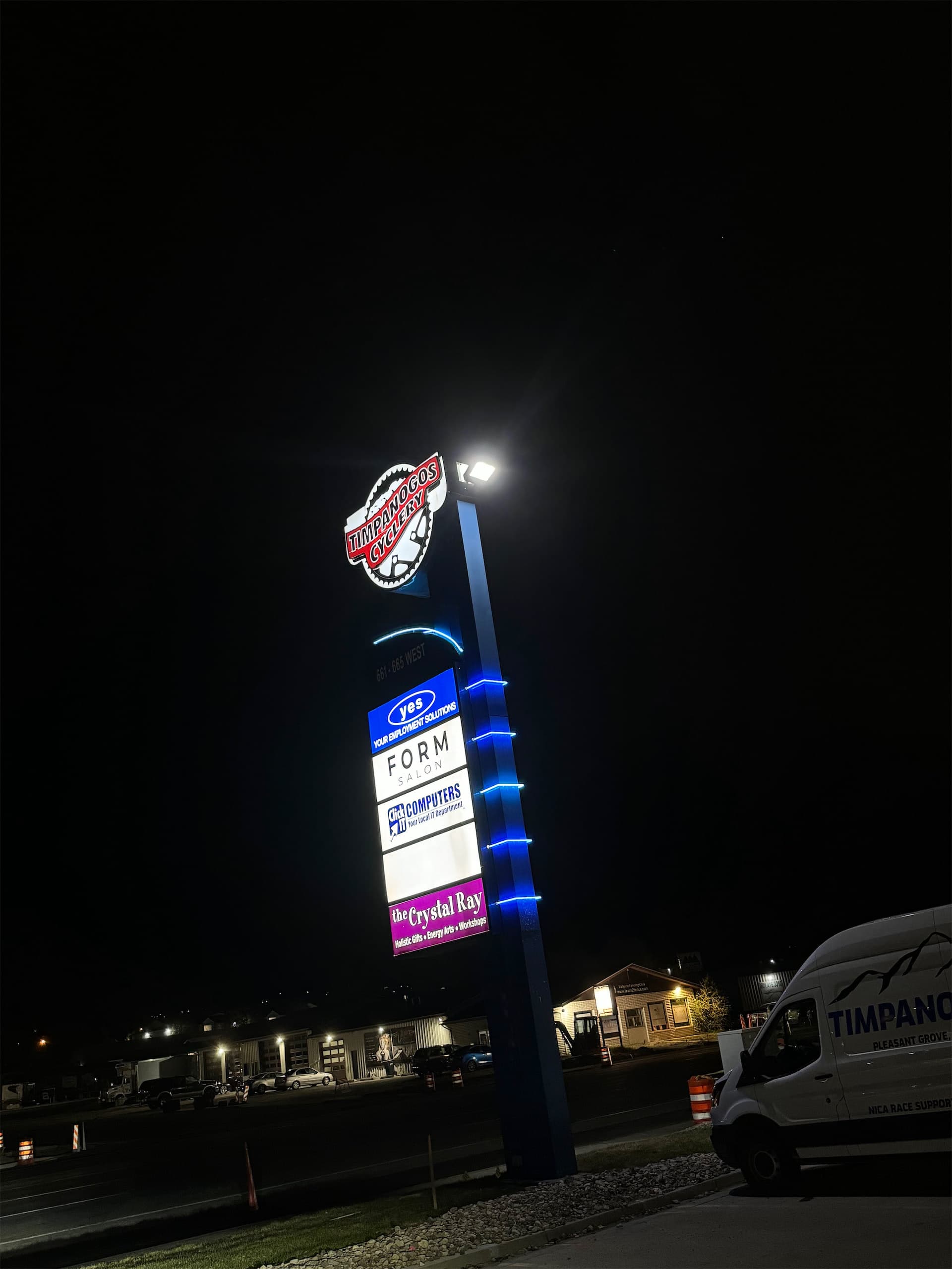Illuminated outdoor pylon sign at night featuring local businesses with blue LED accent lighting.