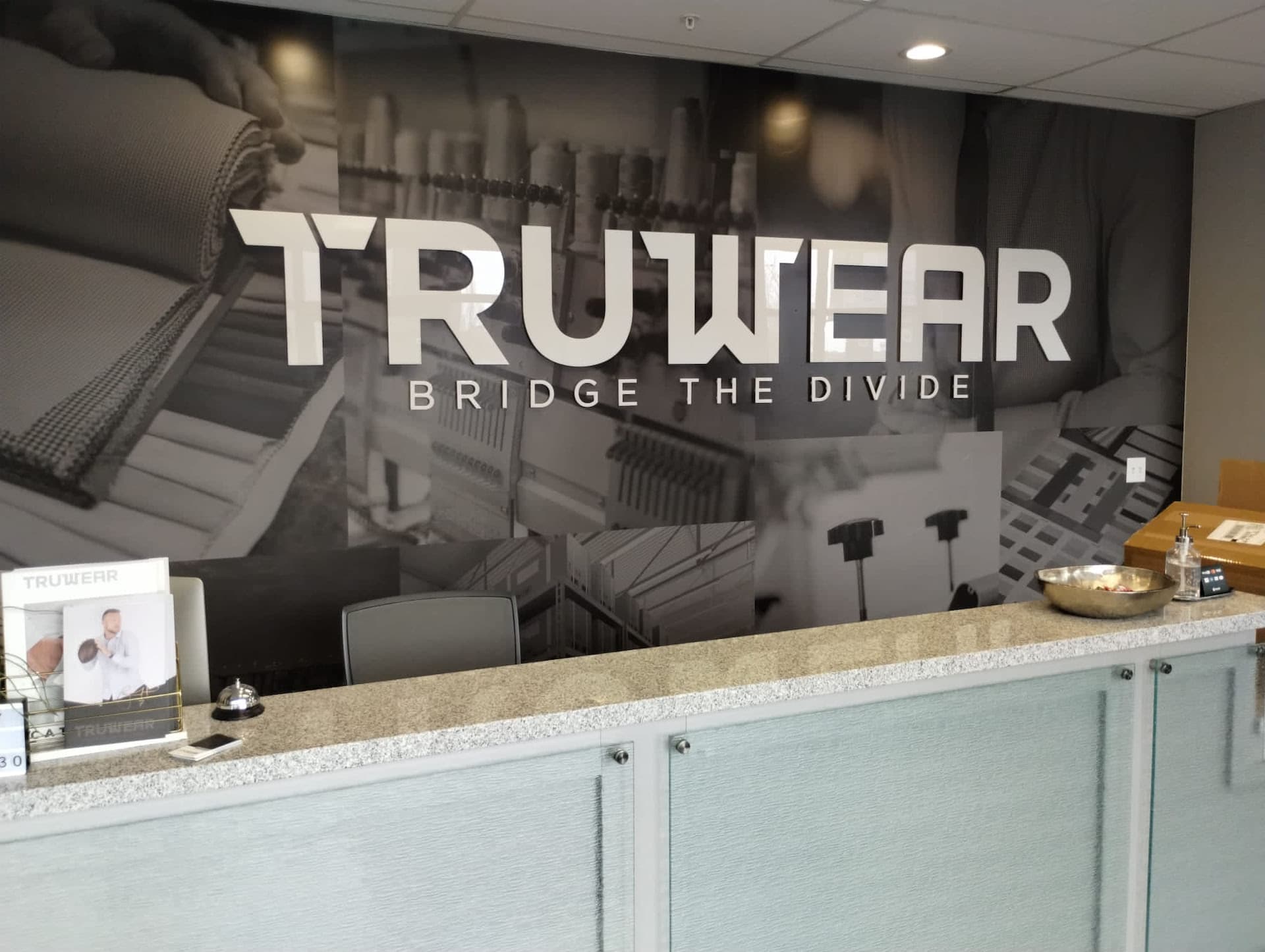 Truwear lobby sign with raised white lettering over a wall mural behind a modern reception desk.