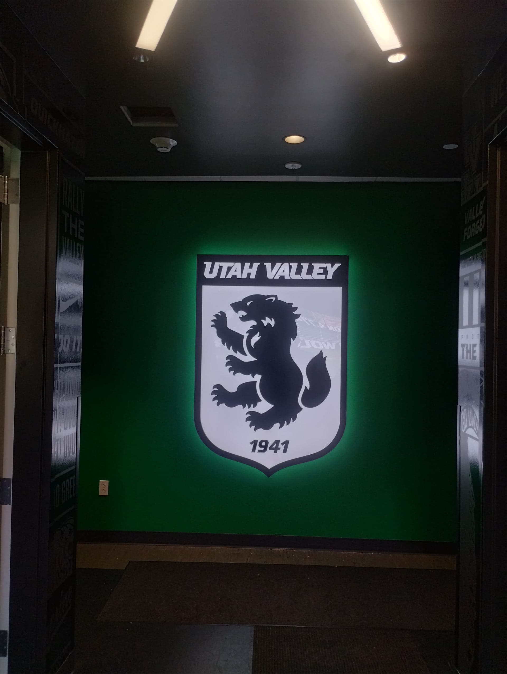 Backlit Utah Valley wolverine crest sign mounted on a dark green wall.