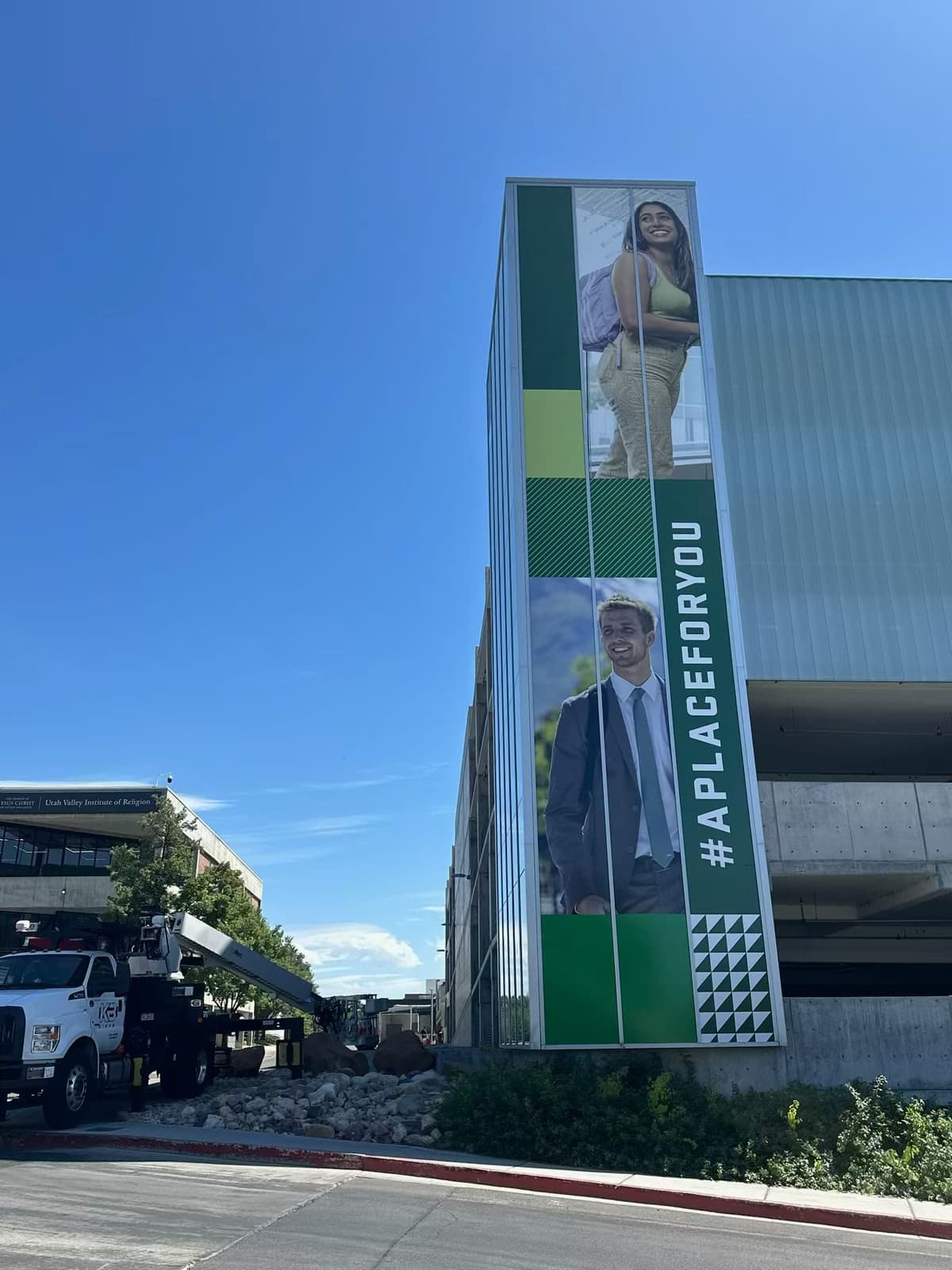 Large green exterior wall graphic on a university building displaying student portraits and the hashtag #APLACEFORYOU.