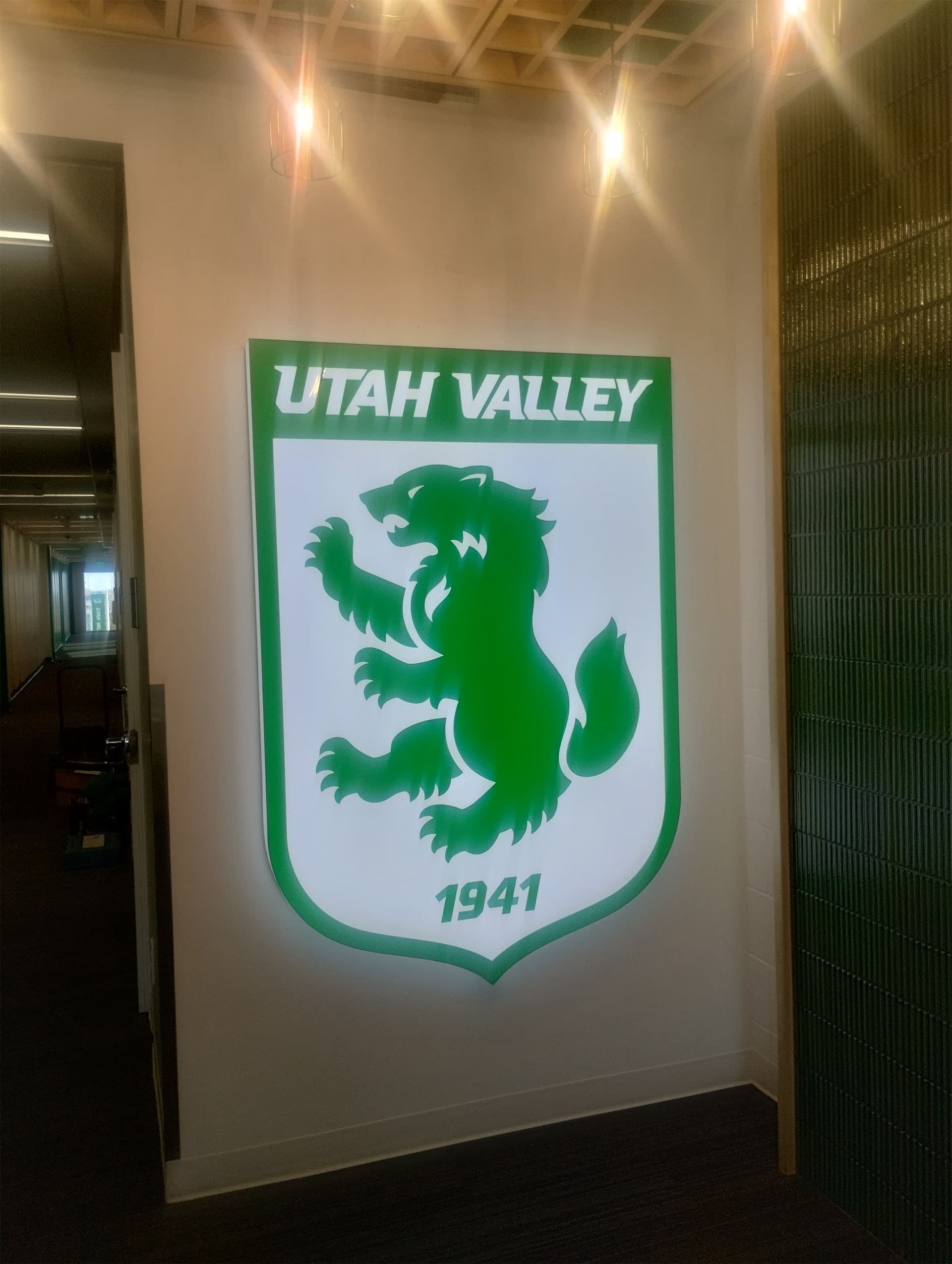 Illuminated Utah Valley University crest sign mounted on an interior wall featuring a green wolverine logo.