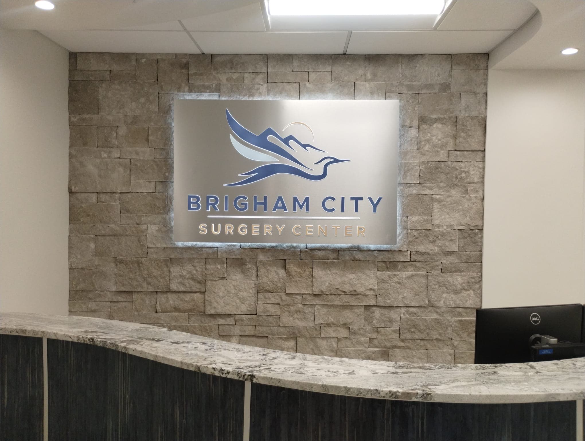 Illuminated brushed metal lobby sign for Brigham City Surgery Center mounted on a stone wall.